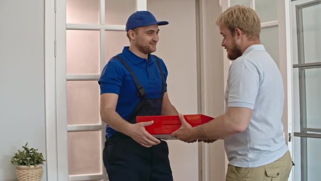 Man opening the door and meeting delivery guy holding two boxes of pizza. He checking his order, giving money to worker and showing thumbs up  