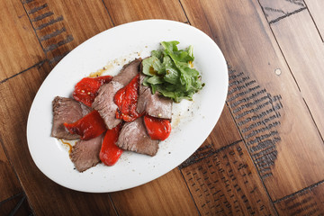 Baked roast beef with rucola and bell pepper in a white oval plate. Wooden background.
