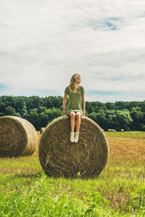 Naklejka na ściany i meble Young blond lady sitting on haystack and looking away on sunny summer day, Badasconytomaj, Hungary