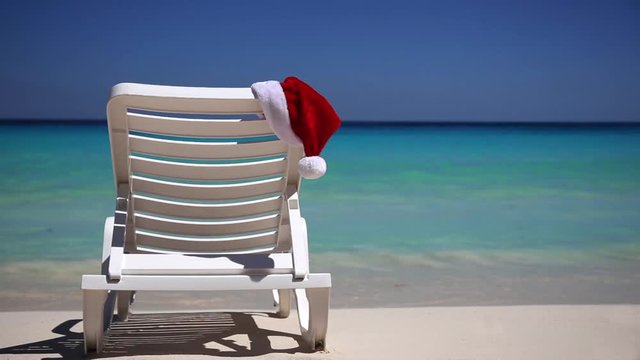 Santa Claus Hat on sunbed near  tropical calm beach with turquoise caribbean sea water and white sand. Christmas vacation concept