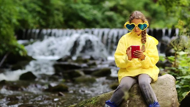 Girl in sunglasses in the shape of hearts with red smartphone sits near small river. Pretty girl playing the game on smartphone. Little girl in yellow jacket with phone sits on big stone near river