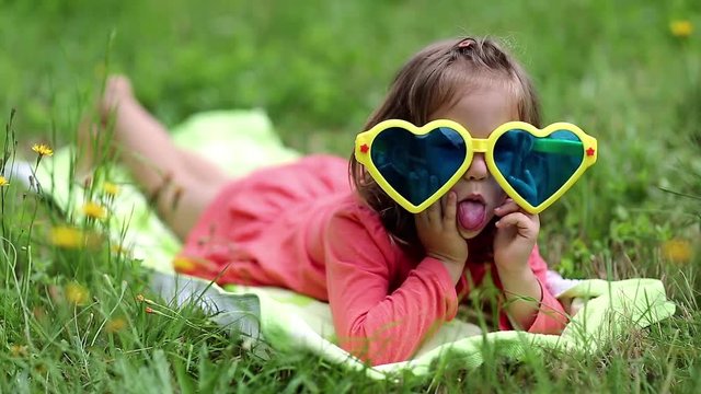 Funny little girl in big sunglasses in the shape of hearts lies on the grass. Beautiful little girl in glasses lies on the green grass and looks at the camera. Funny girl show his tongue
