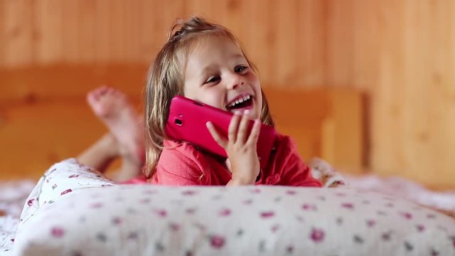 Little girl with smartphone lies on the bed. Funny little girl lies on the pillow and holds in hands red mobile phone, looks at the camera