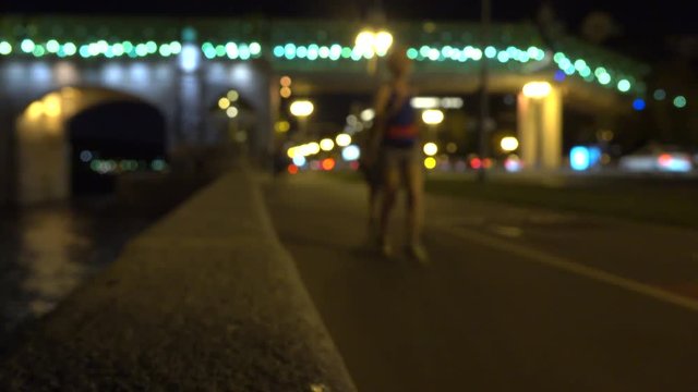 Blurred runner girl and walking people on urban embankment at night. 4K bokeh clip