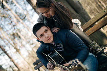 Fototapeta na wymiar Beautiful young couple enjoying outdoor. They siting on a park bench and playing guitar. Autumn day. Haze and back light.