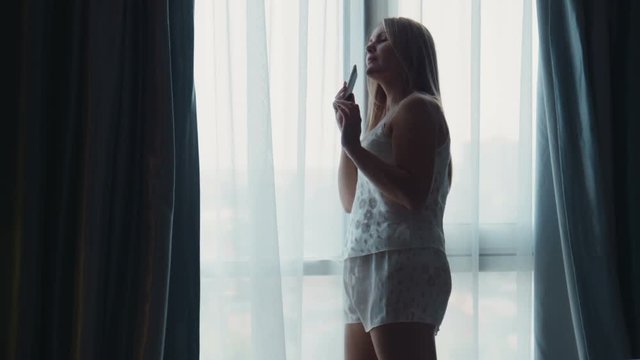 Side view of adult blonde woman in white nighty standing in front of big wide window in sunlight and talking on mobile phone
