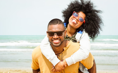 Young afro american couple playing piggyback ride on beach - Cheerful african friends having fun at day with blue ocean background - Concept of lovers happy moments on summer holiday - Vintage filter - Powered by Adobe