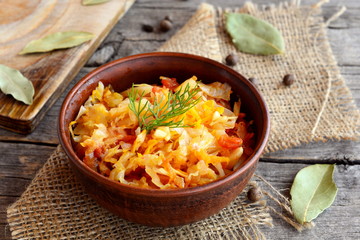 Stewed cabbage in a clay bowl and on old wooden background. Cabbage stewed with carrots, tomatoes and garlic and garnished with a sprig of dill. Vegetable recipe