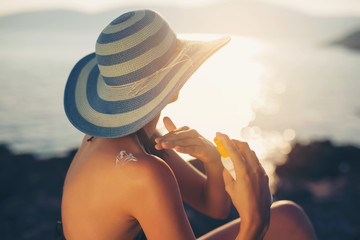 Young woman in sunglasses holding bottle of sunscreen lotion, spraying sunblock cream on shoulder before tanning