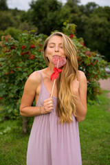 The young girl with a fair long hair dressed in a pink dress holds in hand lollipop in shape heart