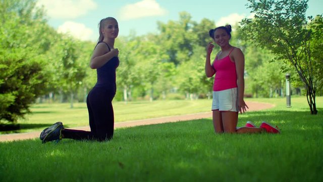 Young woman talking kneeling on green grass in park. Two fit girls in sportswear looking at camera before fitness training outdoor. Multi ethnic friends talks outside. Sporty girls in park