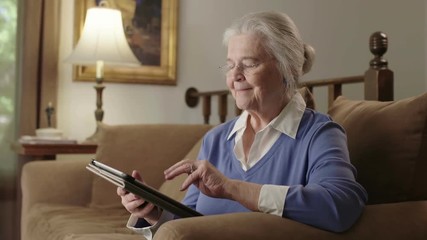 A smiling senior elderly woman uses a computer tablet device on her couch. Shot in 4K UHD. - Powered by Adobe