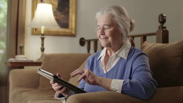 A Smiling Senior Elderly Woman Uses A Computer Tablet Device On Her Couch. Shot In 4K UHD.