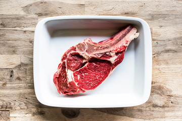 cooking a tasty piece of raw prime rib beef meat in a white porcelain plate on an old oak wood table seen from above ready for the barbecue