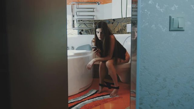 Brunette girl sit on toilet without underwear tapping in smartphone. Bathroom. Apartment