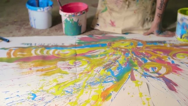 Close up slow motion video of male artist in apron splattering bright  splashes of paint over canvas laid out on floor before him in his workshop studio