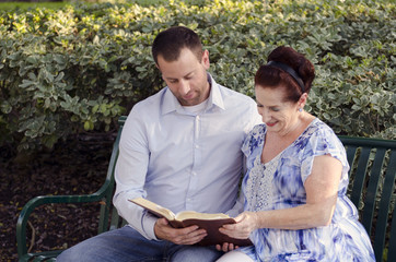 Mother and adult son reading the Bible together in a park sitting on a bench.