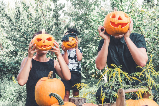 Family holding halloween pumpkin in front of their faces, preparation for party in the garden near Jack-o-Lantern decorations. Father, mother and daughter.