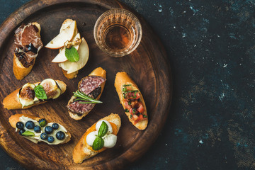 Italian crostini with various toppings and glass of wine on round wooden serving tray over black plywood background, top view, copy space
