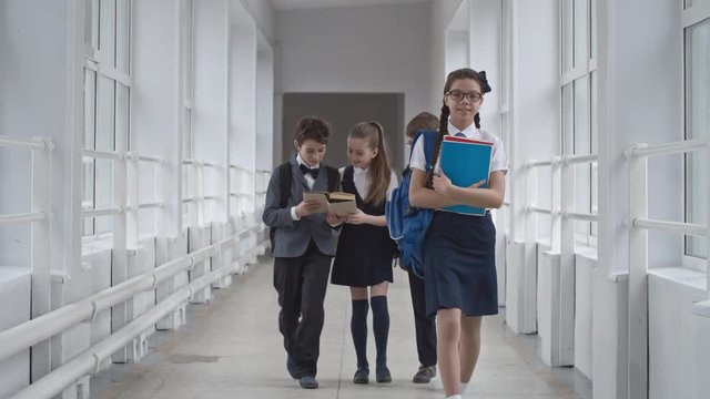 Lockdown of smiling schoolgirl in uniform with course books and glasses walking along school hallway, three other schoolchildren in uniform walking behind smiling and discussing book