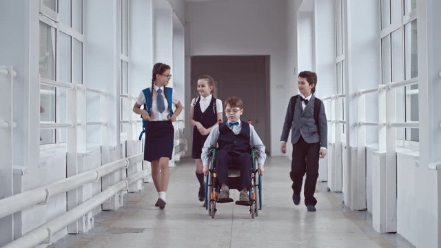 Сoncentrated and sad looking schoolboy in wheelchair wearing uniform and glasses slowly riding along hallway at recess, three other schoolchildren  walking past without noticing him