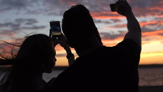 Young happy couple taking photos with smartphones while standing at the beach on sunset