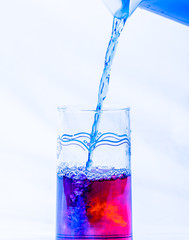 A pitcher of blue liquid being mixed in a glass of red liquid.