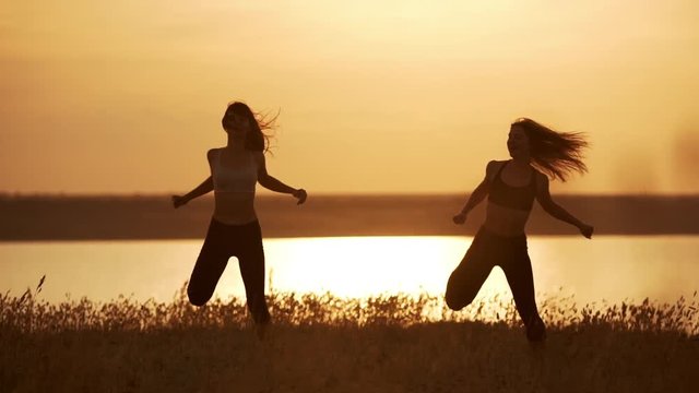 Silhouettes of two beautiful girls dancing zumba in field at sunrise. Slow motion.
