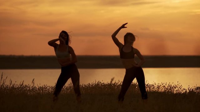 Silhouettes of two beautiful girls dancing zumba in field at sunrise. Slow motion.