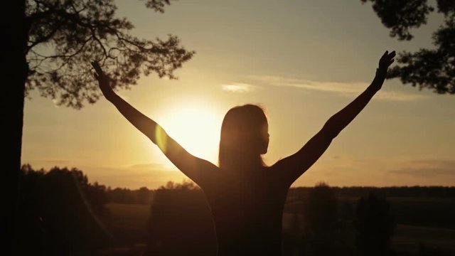 Happy young woman silhouette against bright golden sunset lifts hands up in air. Sunset light, sun lens flares, golden hour. Freedom and happiness concept