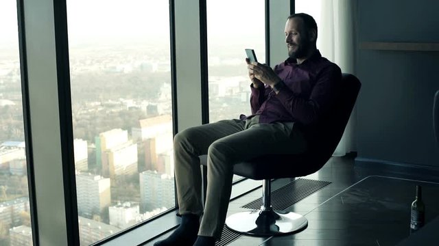 Young, happy man sitting with smartphone on armchair with splendit city view  
