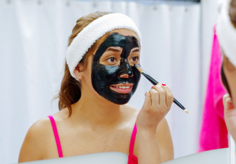 Attractive young woman wearing pink top and white headband, applying black mixture to face using brush, looking in mirror smiling