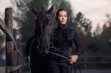 Walking at evening. Portrait of elegant young woman holding her black horse outdoors.