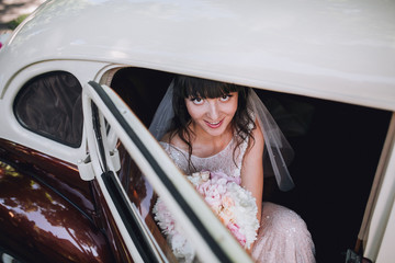 Gorgeous stylish brunette bride posing in retro brown and white limousine car in white dress. Woman...