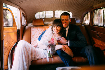 Beautiful happy young bride and groom kissing in retro auto. Embracing near old limousine. Brown color car.