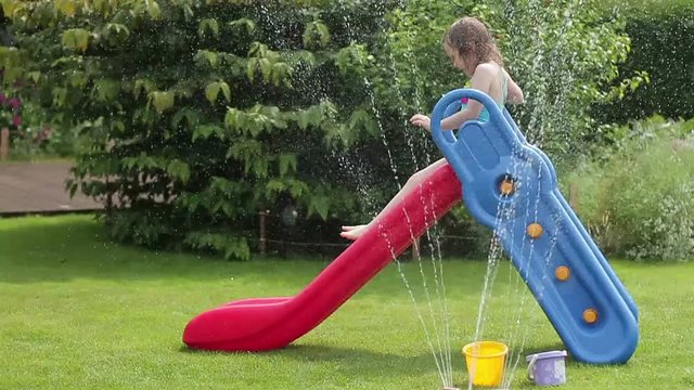 Smiling little girl having fun with garden sprinkler and children's slide in the backyard, slow motion