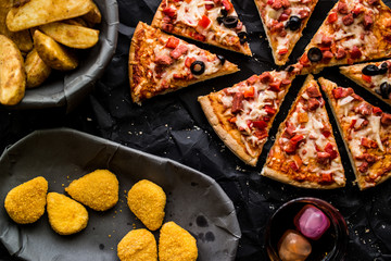 Pizza slices with potato wedges , nuggets and beverage on a black surface.