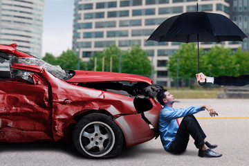 Man with damaged car sits under umbrella