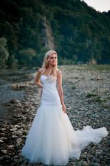 Fototapeta na wymiar Pretty bride in amazing white dress on mountain river's coast