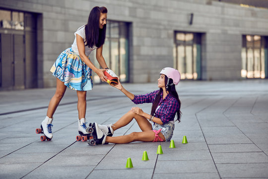 Girl fall down and scratch the leg after rollerblading. Beautiful young woman offers to help protective equipment for the knees. Two girls sports fun. Active lifestyle.
