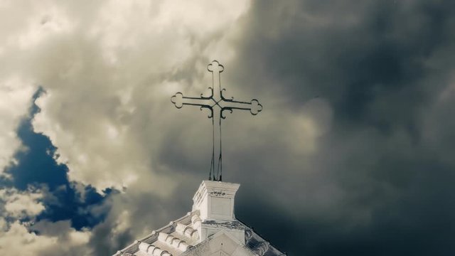 An old stone church under a sky of dark clouds. Detail of the iron cross on the top. Timelapse.
