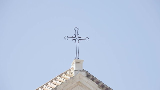 An old stone church over a clear blue sky. Detail of the iron cross on the top.
