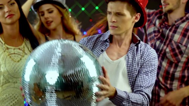 Man with friends keeps disco ball and dancing in nightclub. 4k. Large group people wearing party hat have fun and dance in nightclub.