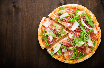 Sliced pizza with prosciutto (parma ham), arugula (salad rocket) and parmesan on dark wooden background top view. Italian cuisine. Space for text. - obrazy, fototapety, plakaty