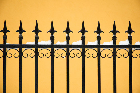 old fashioned spike fence against yellow wall, some snow