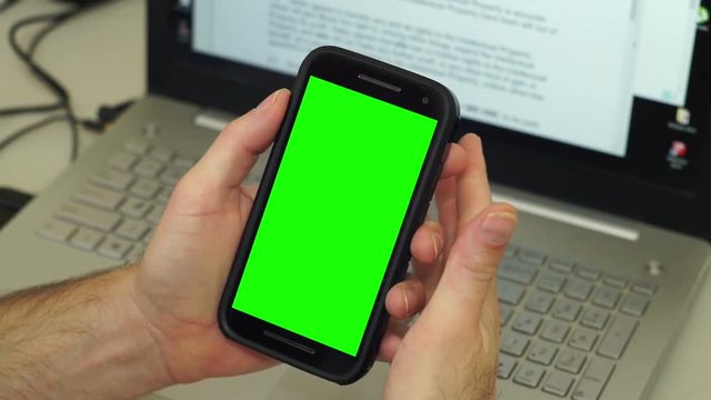Anonymous man in front of his computer makes gestures with his hands holding a mobile phone or cellphone with a greenscreen or chroma key screen seems angry or annoyed with what he sees.