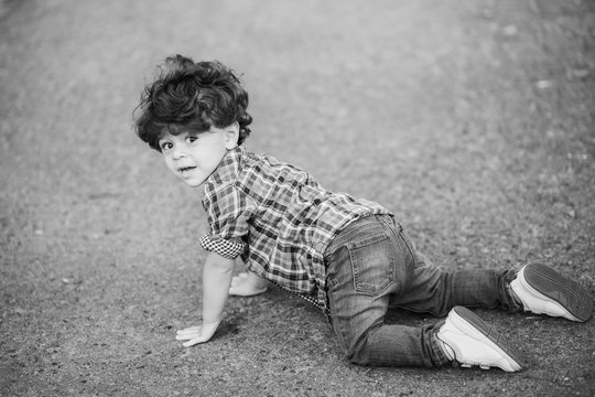 little cute toddler boy crawling outside. Caucasian child dressed in casual clothing: denim jeans, shirt and sneakers. Portrait of kid with beautiful dark curly hair. Horizontal black and white image.