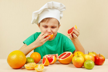 Little boy in chefs hat eat fresh acidic grapefruit at the table with fruits