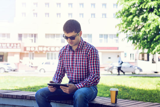 man in  shirt and jeans  sunglasses, video looks on the tablet corresponds to the social networks, in the summer  the bench, the concept of  businessman  vacation. City lifestyle.  the street in the