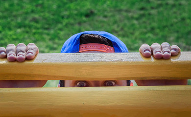 Looking for something concept. Man's eyes between laths of bench in park and hands on top.